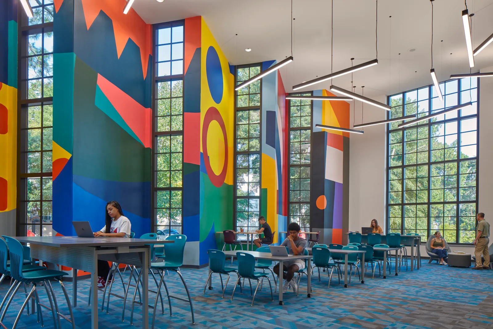 A handful of students sit at desks in a large room with tall walls with murals and glass.