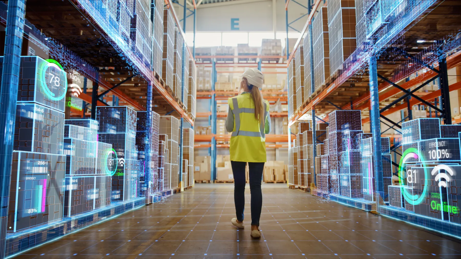 A woman in a yellow safety vest walks in a warehouse with boxes showcasing digital readings.