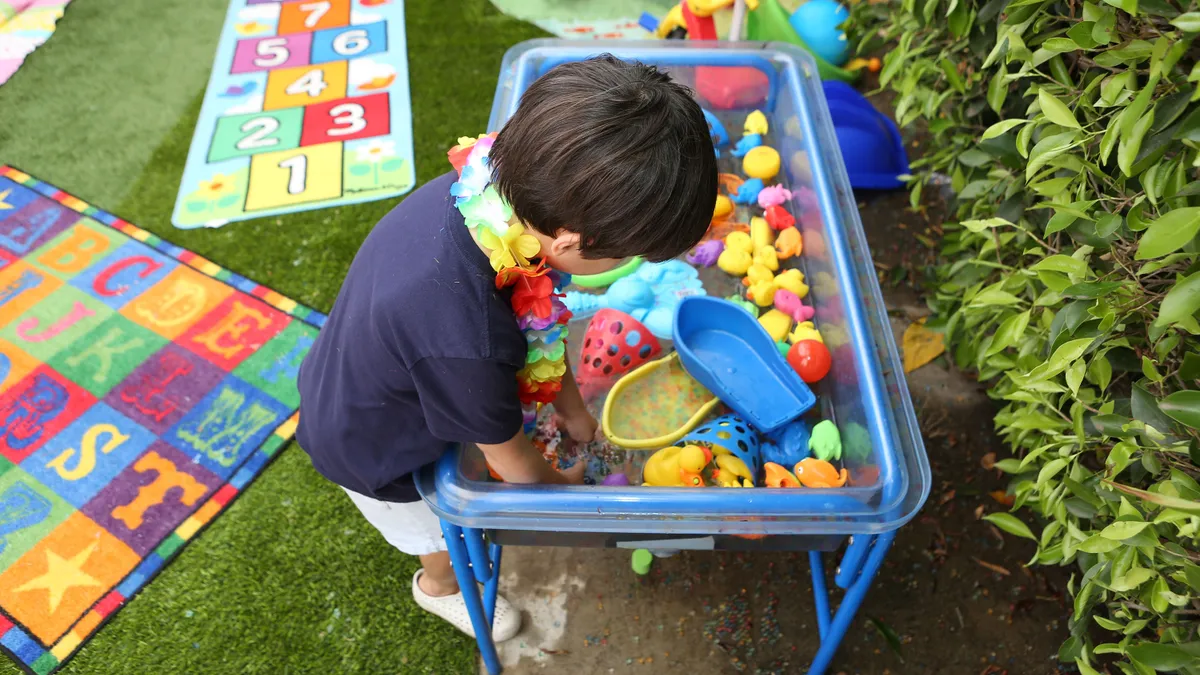 A small child reaches into a blue plastic bin of brightly colored toys on a sunny day during a kindergarten readiness summer learning program.