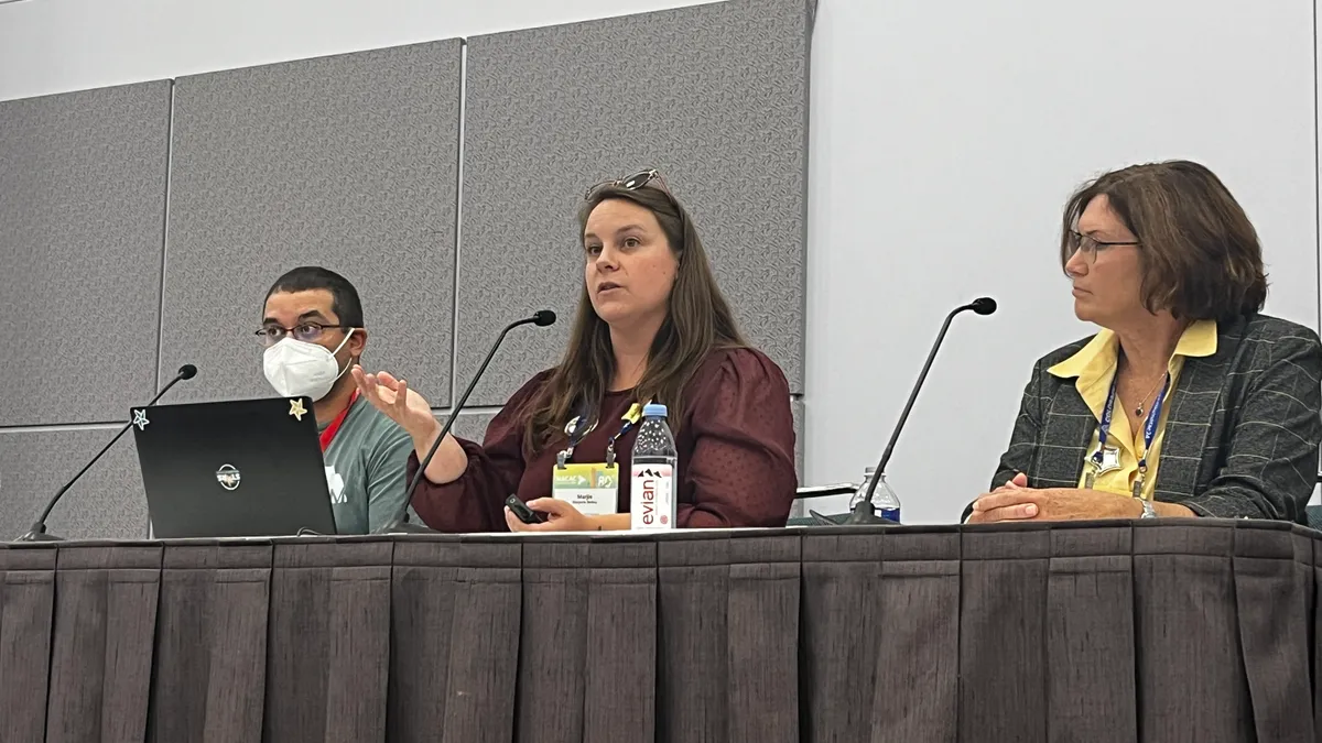 Three people, one of them wearing a N95 mask, sit at an on-stage table.