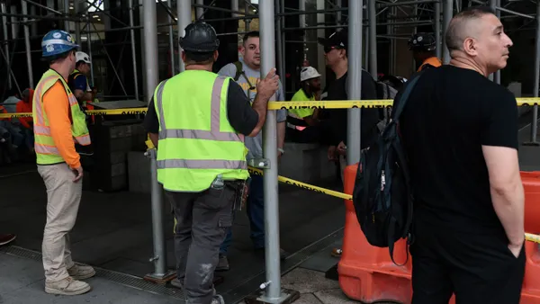 A group of construction workers and citizens stand and sit under construction scaffolding.