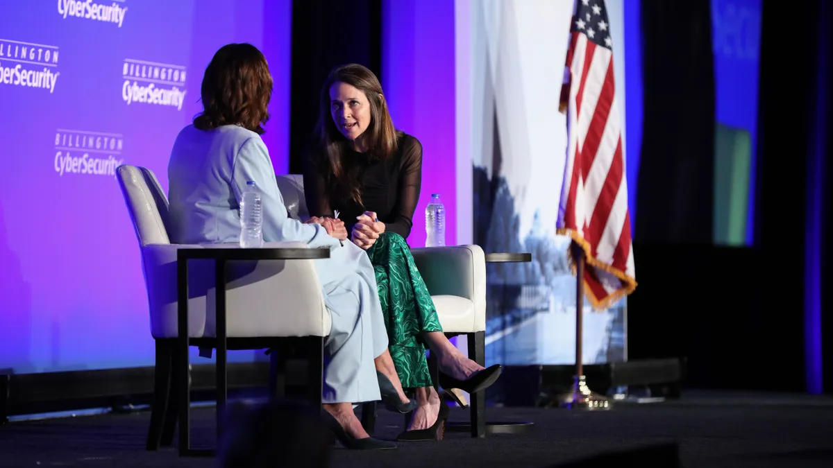 Two people sitting on a stage during a conference, with a U.S. flag in the background.