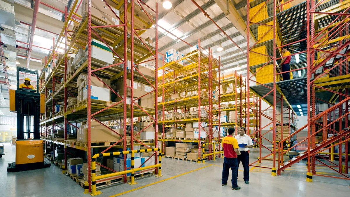 Workers talk while a man uses a piece of equipment to reach higher levels of a storage area.