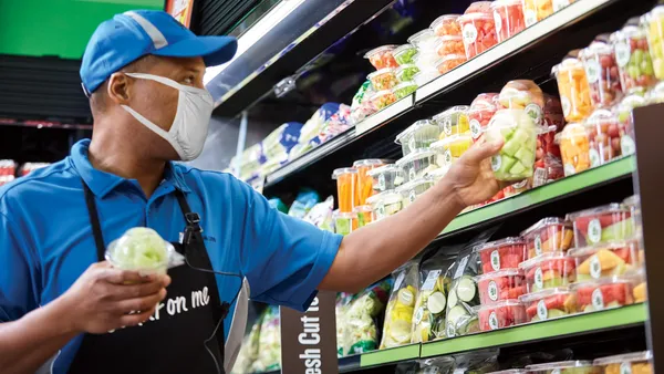 A worker wearing a face mask, cap and apron places a package of precut fruit on a grocery store shelf.