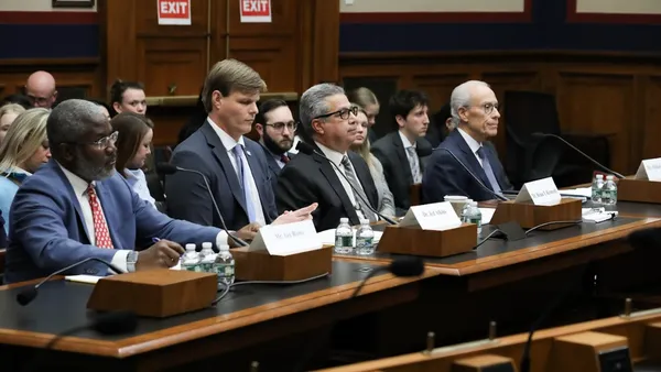 Four people sit at a table. On the table are microphones. People seated in rows are behind them.