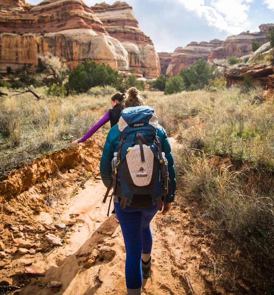 Two people hiking single file toward massive rock formations, seen from the back, with a backpack on the person behind.