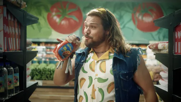 A man with long hair wearing a denim vest with a short-sleeve button-up shirt with a print of chilis and tacos holds a bottle of Ortega sauce as if to drink it.