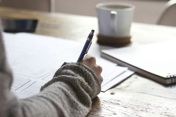 Person writing on brown wooden table near white ceramic mug