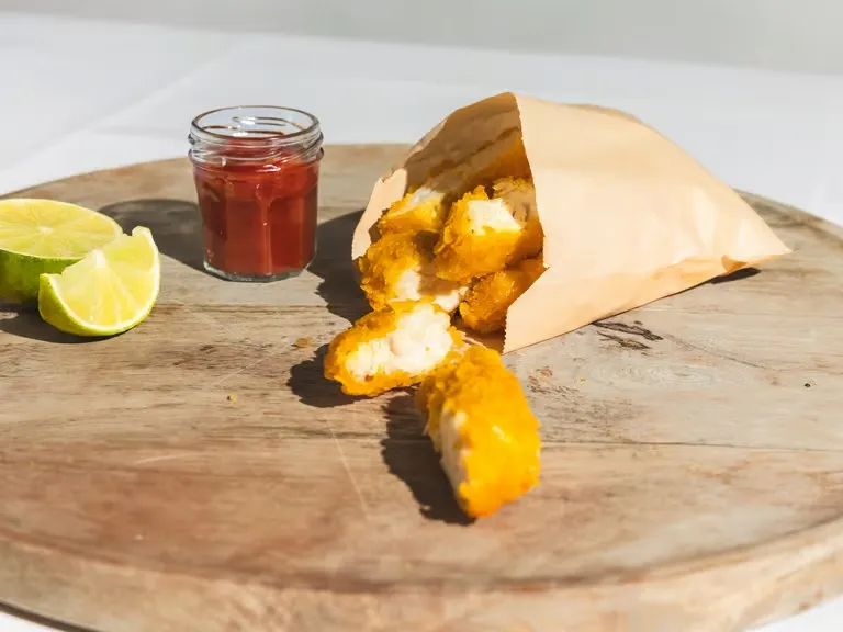 A small paper foodservice bag holds fried chicken nuggets and is positioned next to a pot of ketchup.