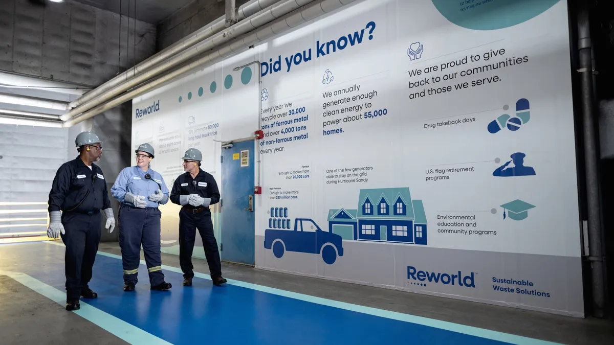 Three people wearing hard hats and work gear walk down a hallway next to a sign touting Reworld's accomplishments.