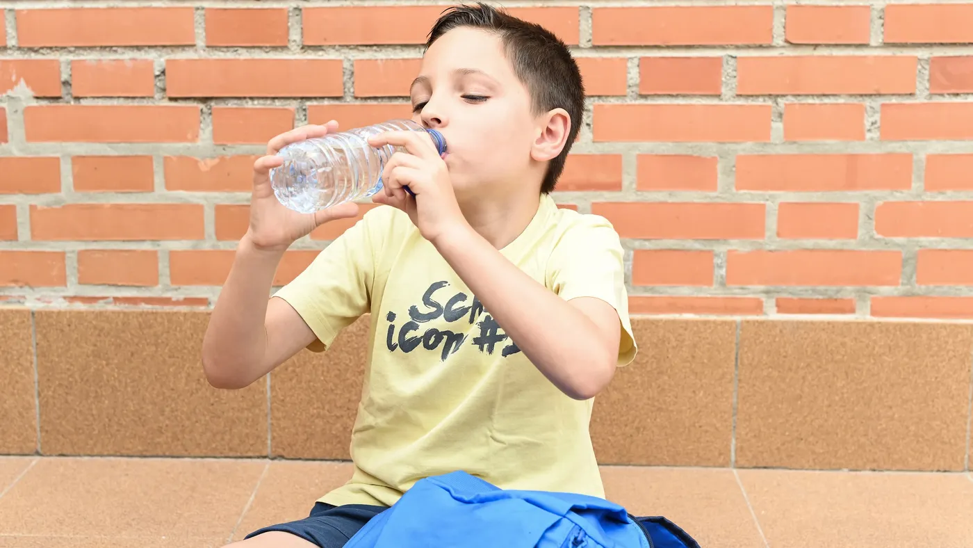 A child sitting next to a backpack in front of a brick wall drinks from a plastic water bottle.