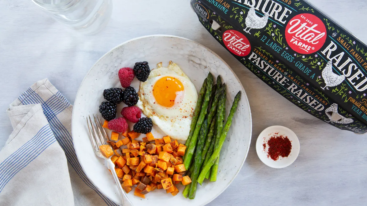 A breakfast plate with sweet potatoes, berries, asparagus and a fried egg. A glass of water and a carton of eggs are also shown.