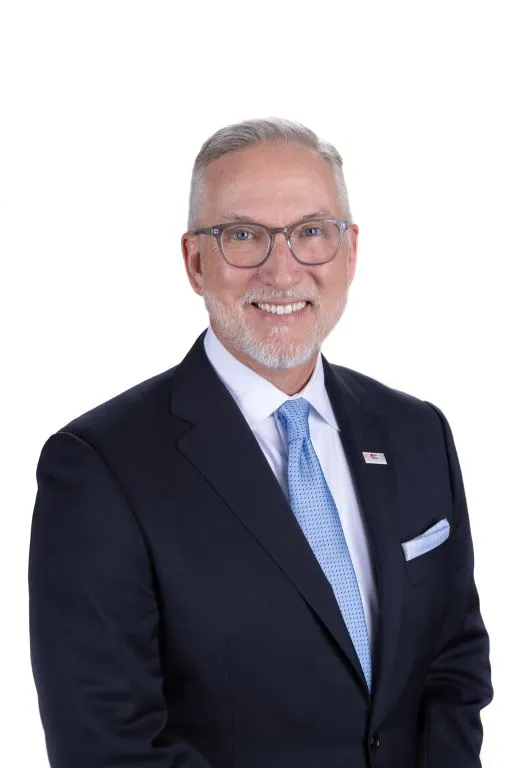A smiling white man in a dark suite with a light blue tie, white shirt and white facial hair.