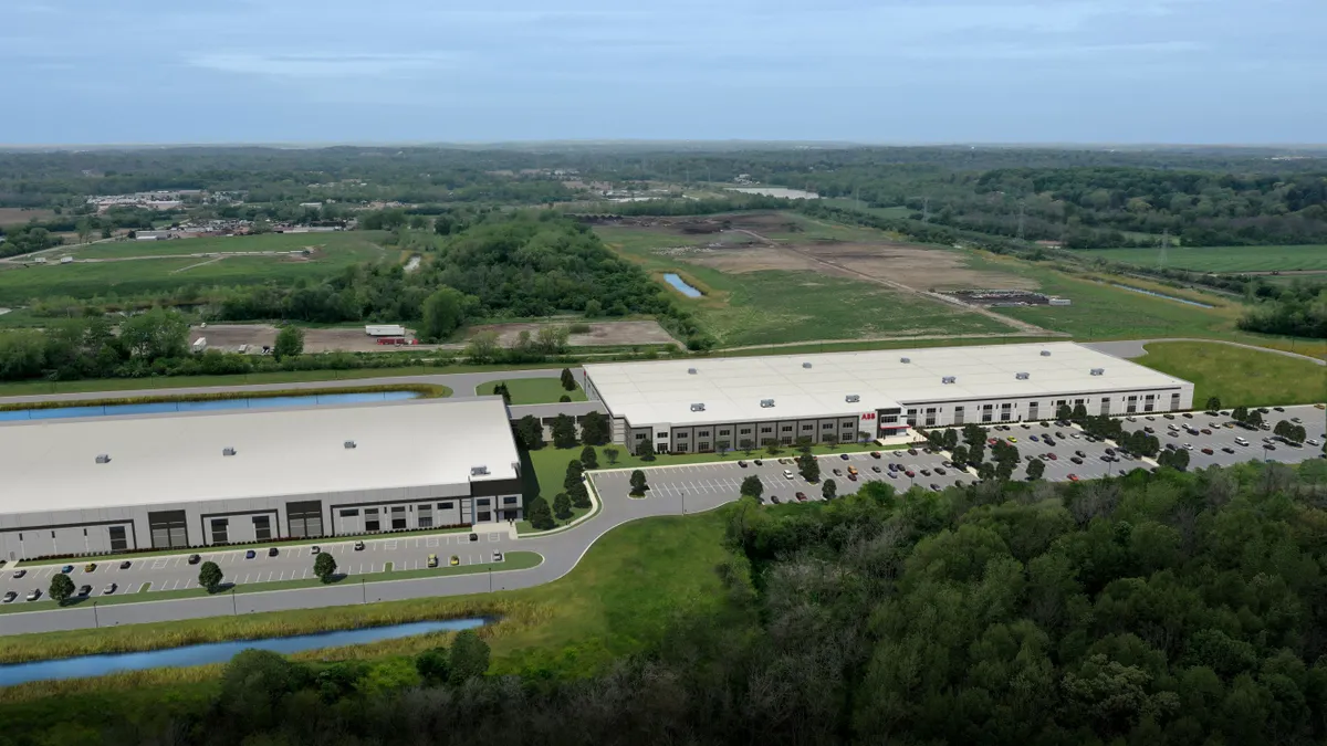 Two white buildings with light gray parking lots on the front, with various colors of cars, and surrounded by green trees and grass.