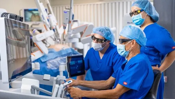 Three clinicians in surgical garb view the screen at the console of Medtronic's Hugo robotic surgery system.