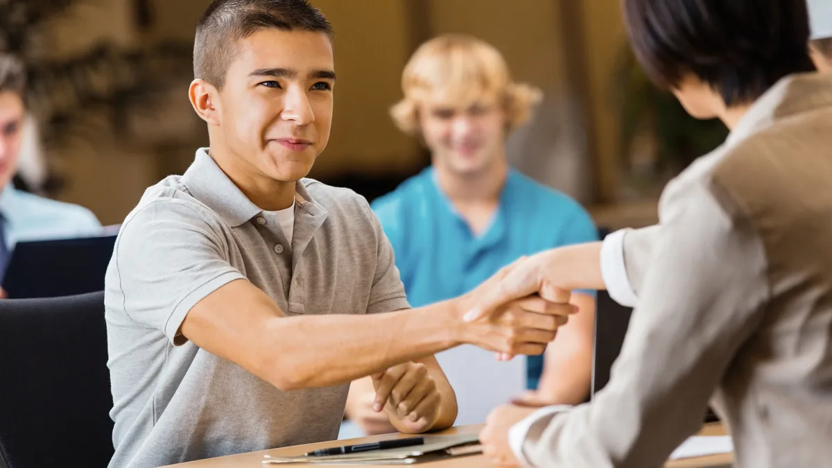 A boy high school student shakes a woman professional's hand across a tabletop during a career fair interview.