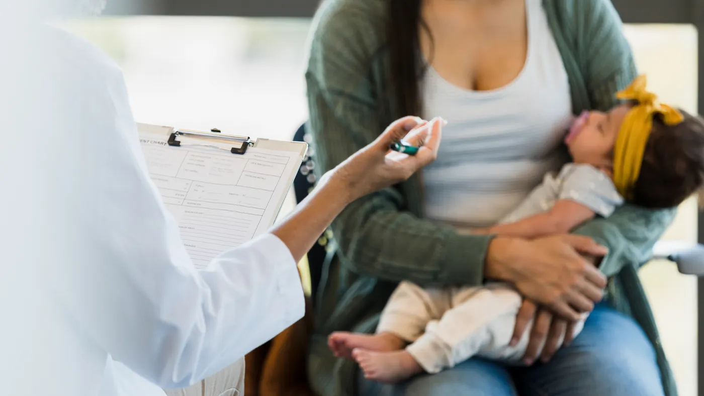 A doctor holding a clipboard speaks to a women with a newborn baby.