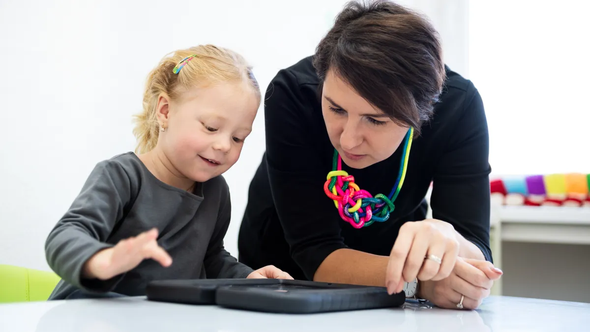 A student and adult sit at a table. The student is touching a tablet on the table