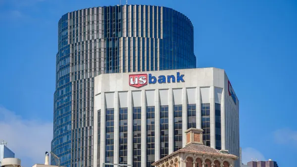 A tall modern office building with a white facade and the "US Bank" logo at the top, positioned in front of a taller cylindrical glass skyscraper.