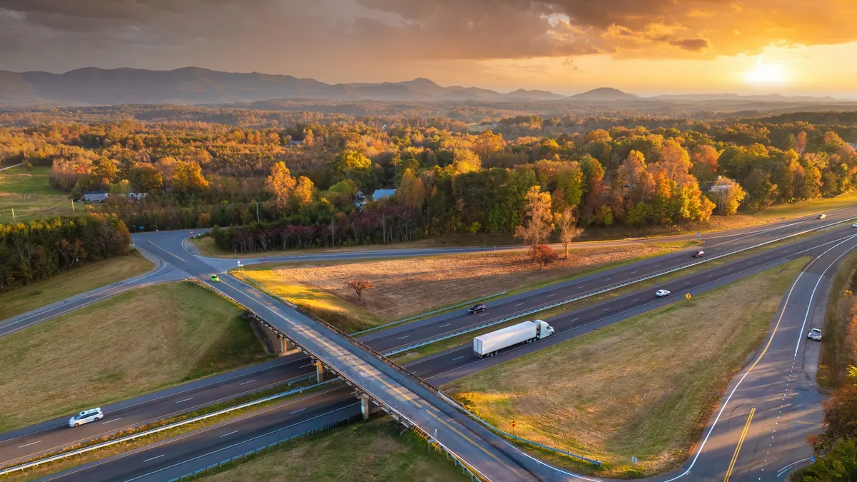 A highway overpass junction with traffic, including a tractor-trailer and passenger vehicles, at sunset.