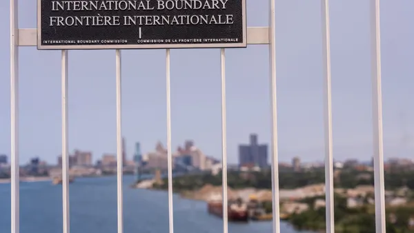 Metal sign that says "United States Etats-Unis Canada International Boundary Frontiere Internationale" on metal bars with body of water and city skyline behind it.