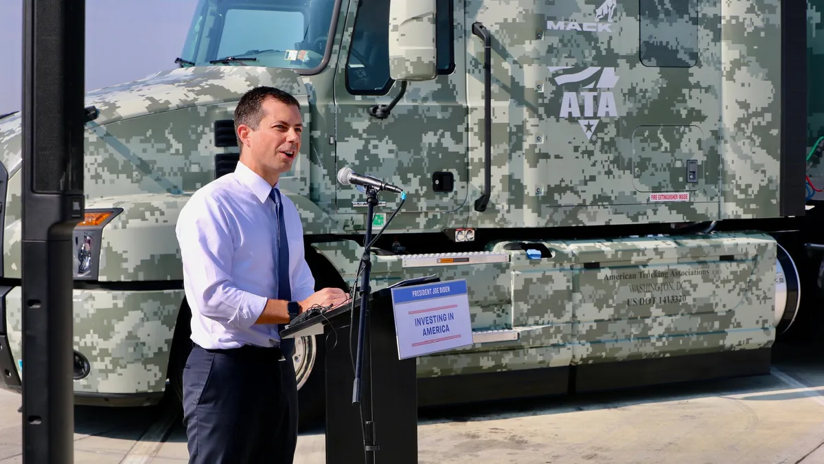 Transportation Secretary Pete Buttigieg announces truck parking and other highway safety grants in front of a camouflage ATA Workforce Heroes truck.