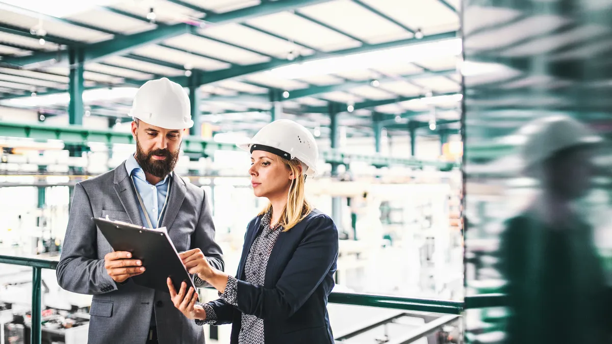 Two employees in a warehouse wearing hard hats, reviewing paperwork.