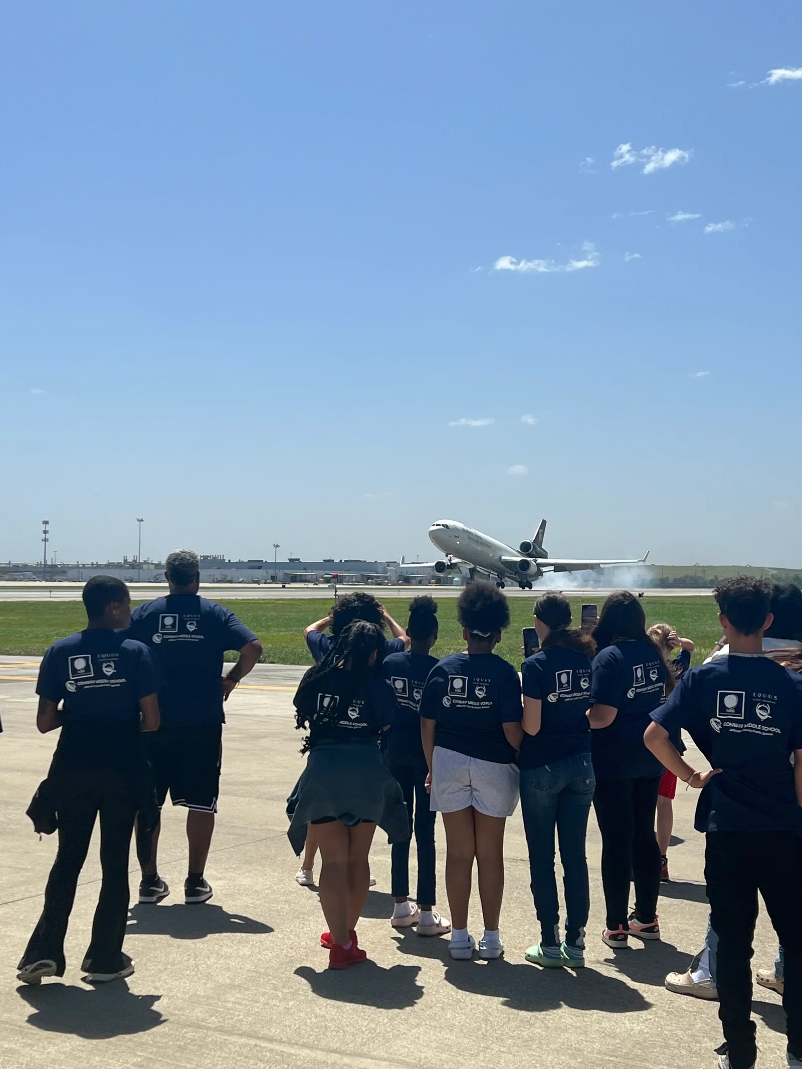 A group of students, wearing the same dark blue T-shirts, stand outside at an airport, watching a plane take off.