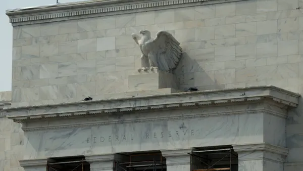 A white marble American eagle sculpture sits atop a white marble building entrance, with the words "Federal Reserve" seen etched into the marble.