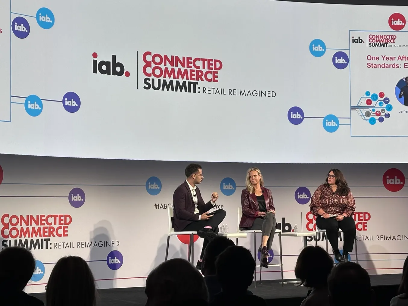 Three individuals on stage sitting on chairs talking to each other. Background is a white screen that reads "IAB Connected Commerce Summit."