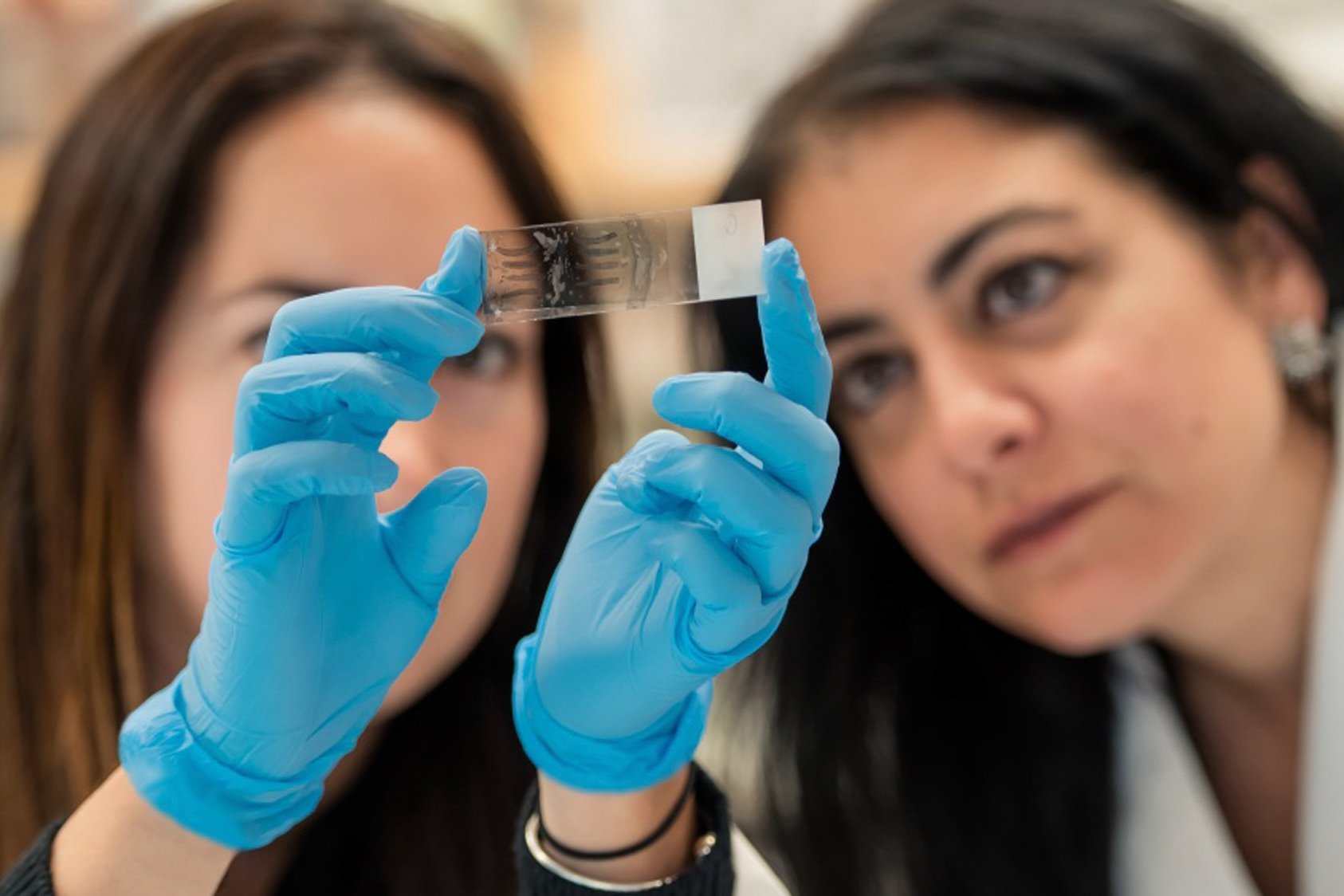 Two people look at a microscope slide being held up by a pair of gloved hands.