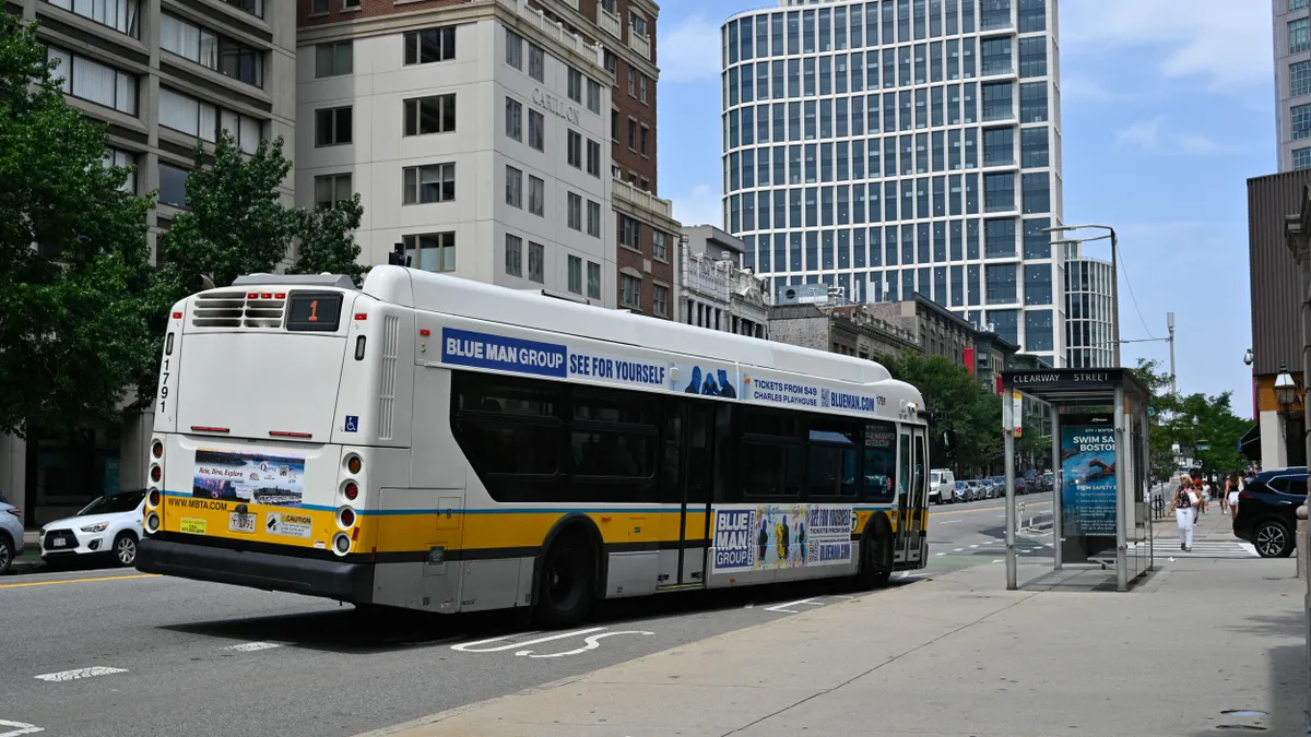 A white transit bus with advertising is seen from the rear as it makes a stop in a bus lane next to a bus shelter on a city street.