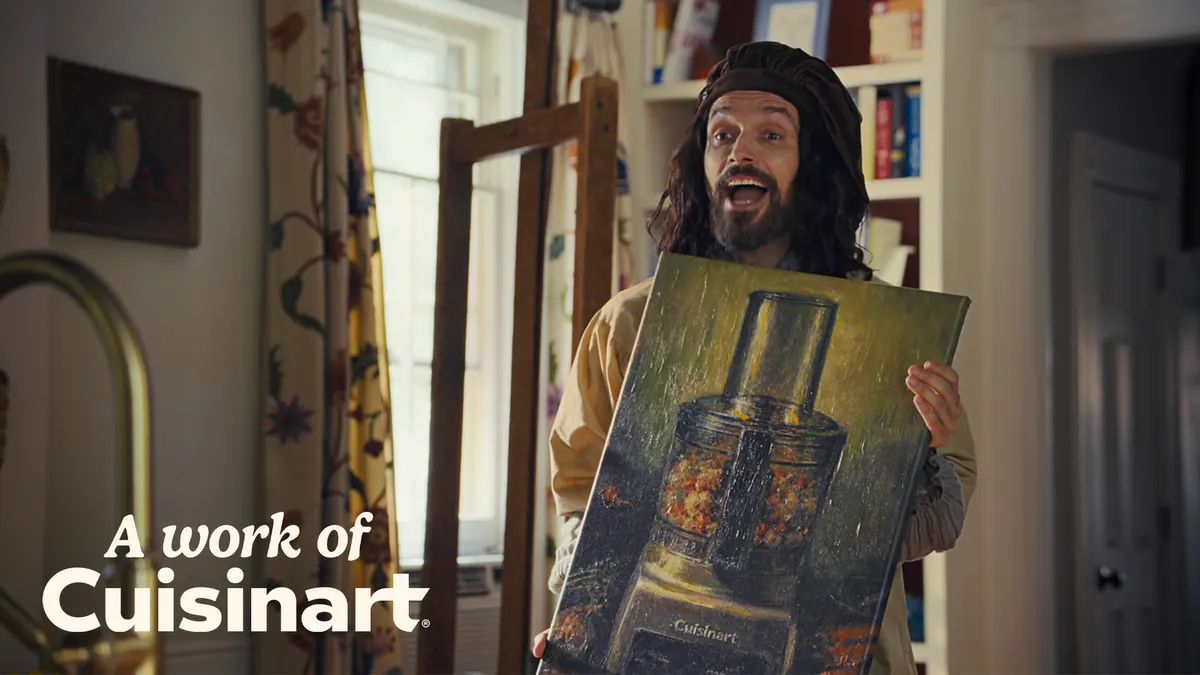 A Baroque period-styled artist holds up an oil painting of a Cuisinart product in a modern kitchen