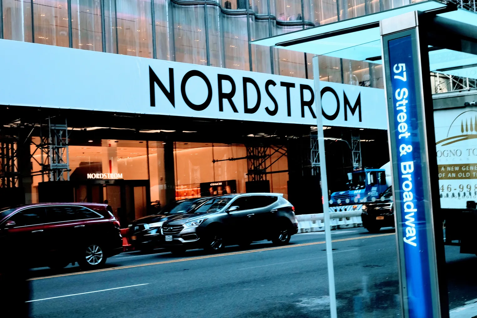 A bus stop with a bright blue sign that reads "57th Street and Broadway" sits across the street from a Nordstrom store.