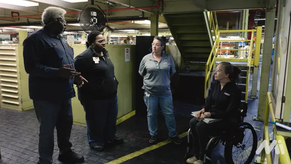 Four people wearing safety glasses and dark clothes, three of which are standing and one in a wheelchair inside a factory.
