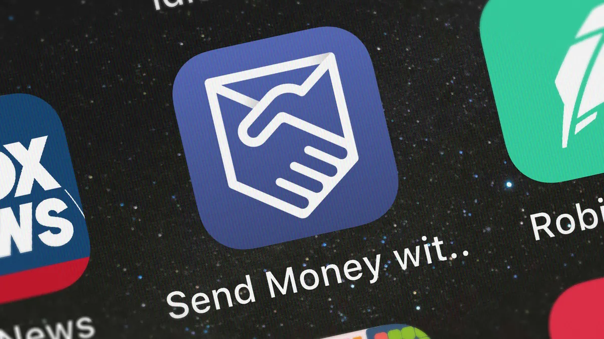 Close-up of a smartphone screen showing an app icon with a blue background and a handshake logo inside a stylized envelope. The text below reads "Send Money wit..".