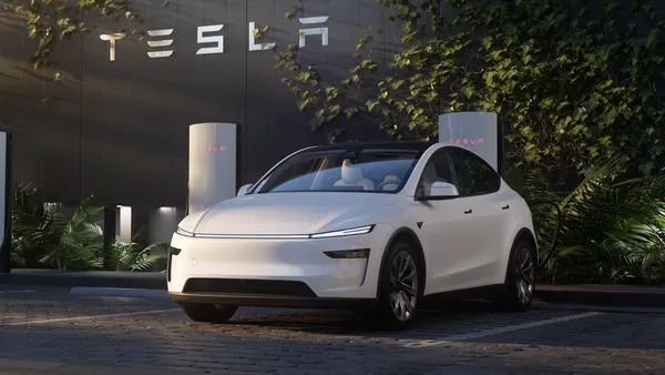 A white Tesla Model Y parked at a Supercharger with a Tesla logo shown on a building wall in the background.