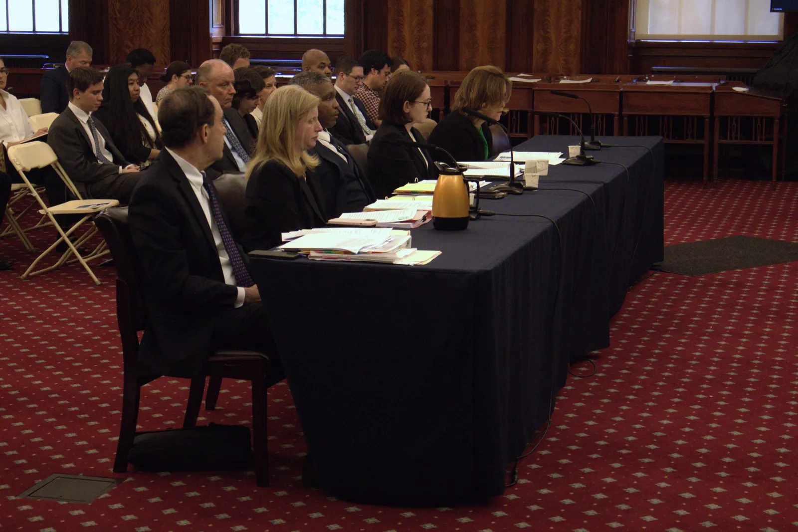 Several people dressed formally sit at a table in front of microphones in a plush carpeted room with wood paneling.