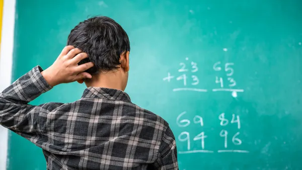 A student with their back to the camera faces a green chalk board. On the board are math problems. The student has their hand on their head.