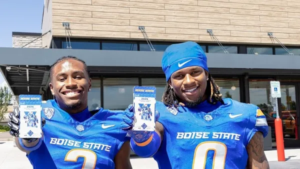 A photo of two people dressed in blue football uniforms holding trading cards and standing outside a store. A sign on the store says Jacksons while their uniforms say Boise State, with one having the number 3 and the other having the number 0.