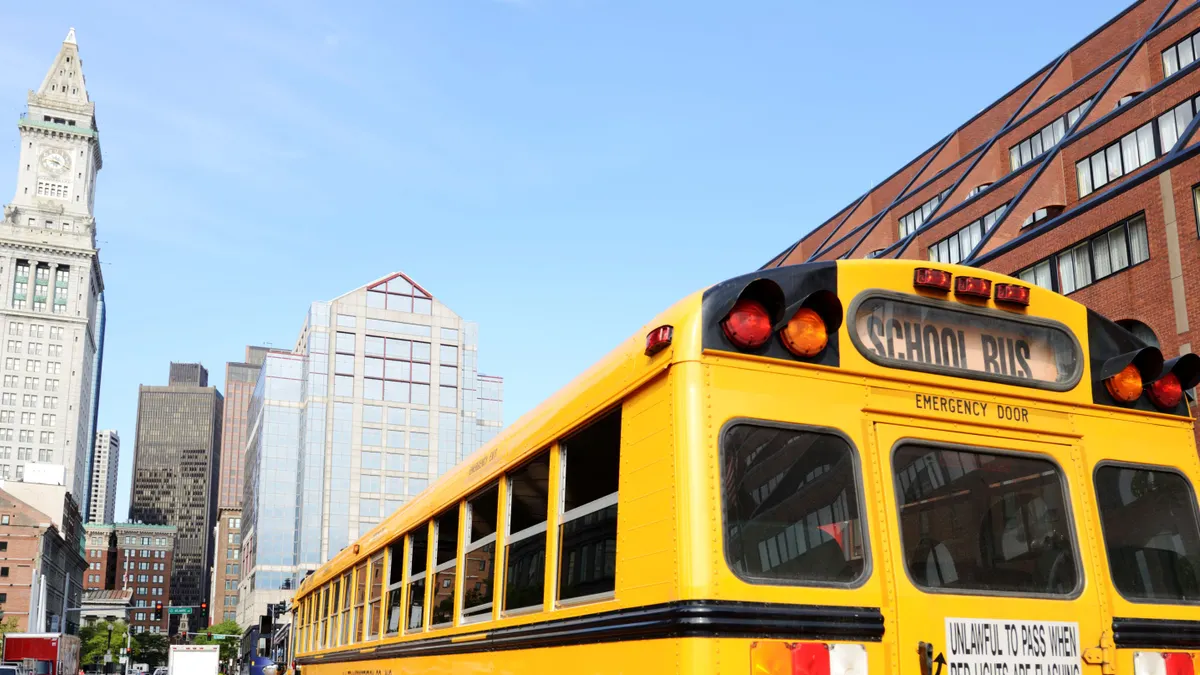 A yellow school bus drives along a road with tall skyscrapers ahead.
