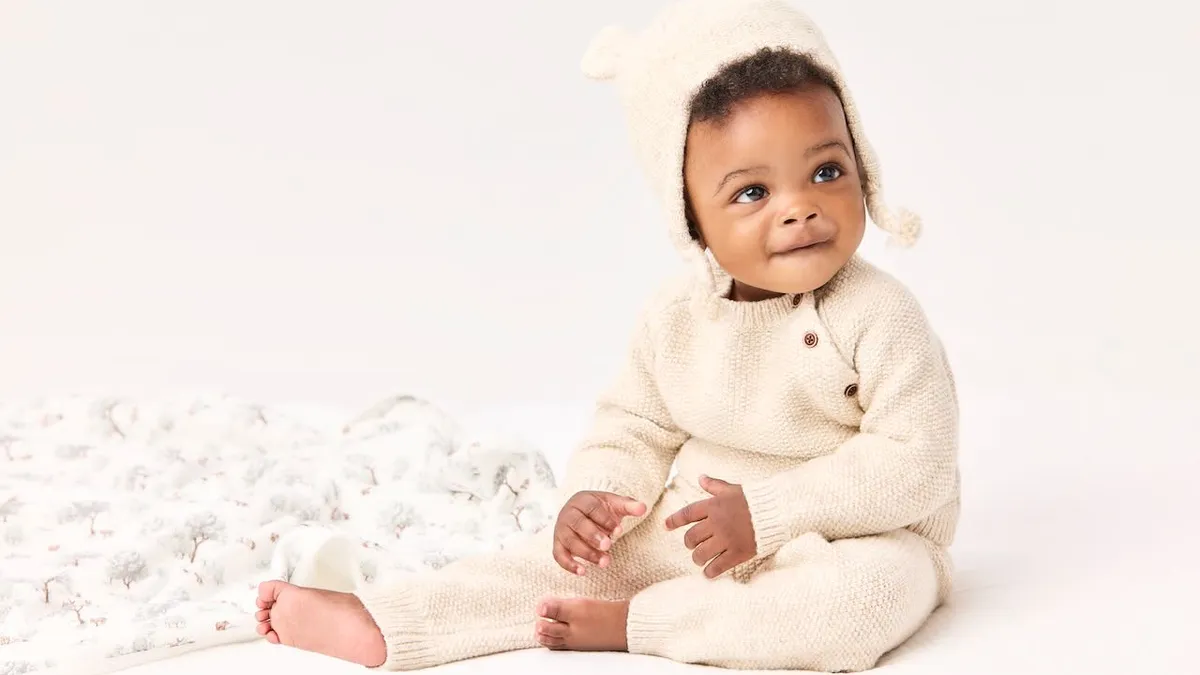 A baby dressed in white knit onesie and cap sits next to a white blanket, looking up.