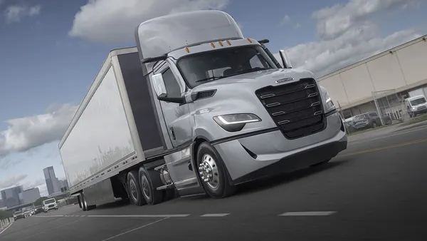 A Freightliner Cascadia's fifth generation tractor-trailer on a highway with city buildings and traffic in the background.