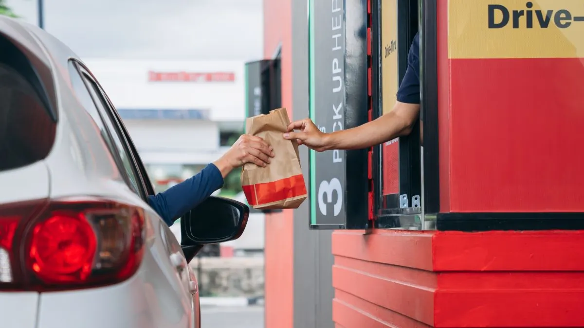 An arm reaches out of a gray car to collect a bag of food while an employee with blue gloves hands off the bag.