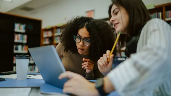 Two students look at a laptop together while in a library.