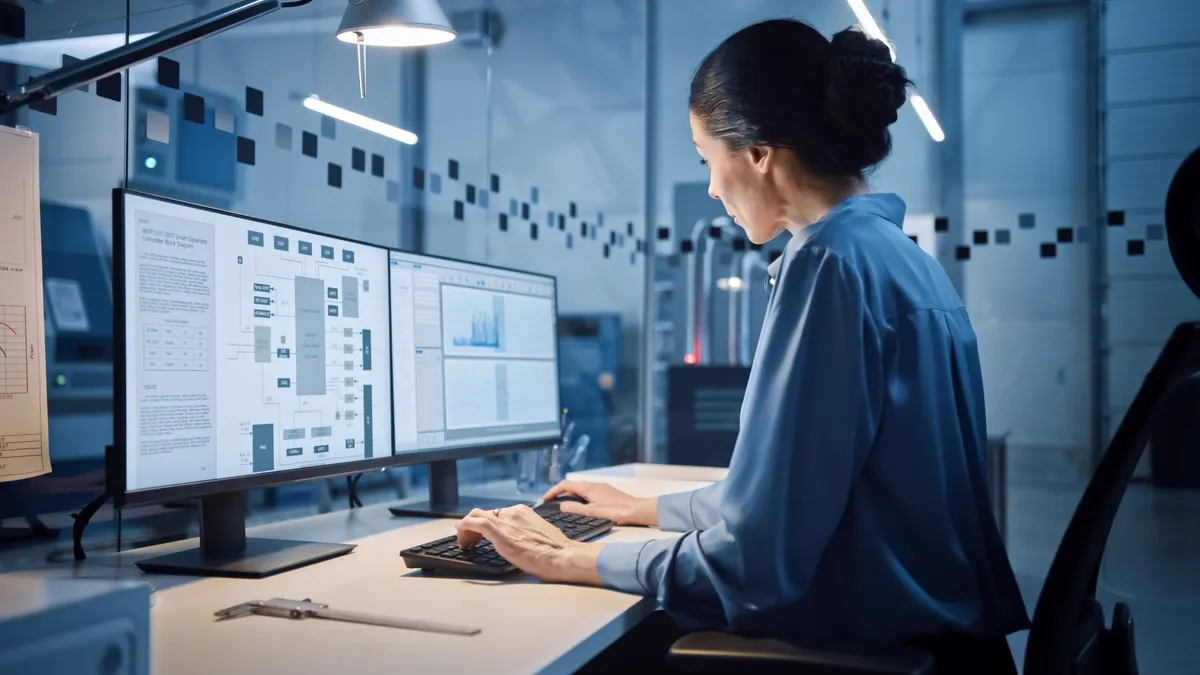 A woman sits in an office, working at a computer with dual screens.