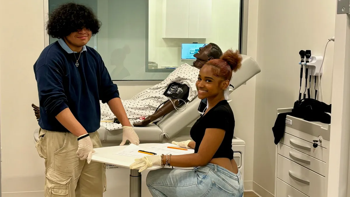 Two students are in a room with a mannequin laying in a bed. The students are looking at the camera.