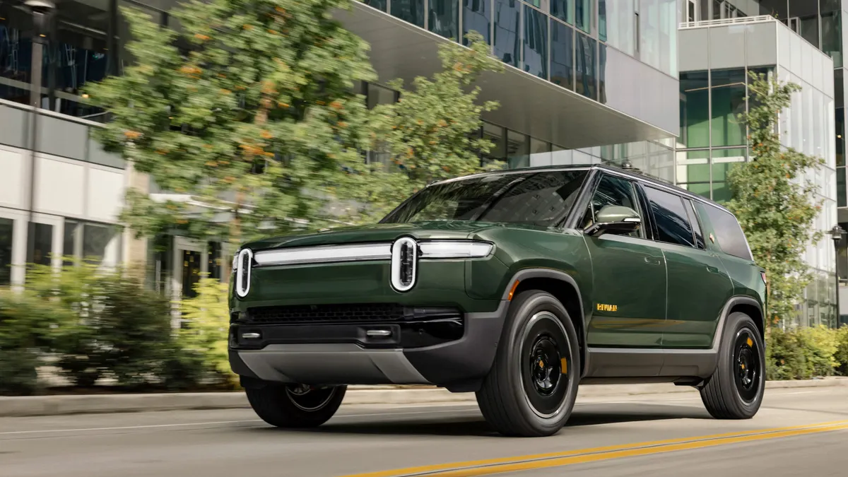 A dark green Rivian R1S electric SUV driving on a city street with buildings in the background.