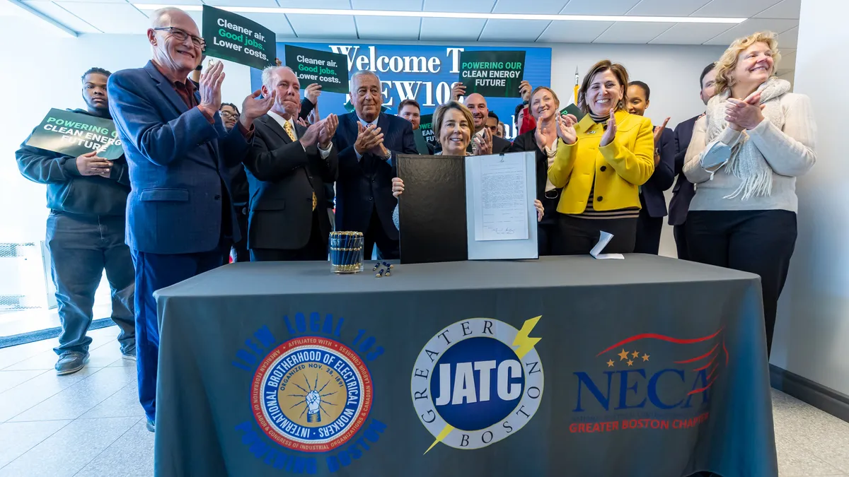 Maura Healey, seated at a table, holds up a climate law, surrounded by a group of people clapping.