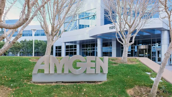 The exterior of Amgen's office building in South San Francisco, with a sculpted company sign in the foreground and leafless trees on a grassy lawn.